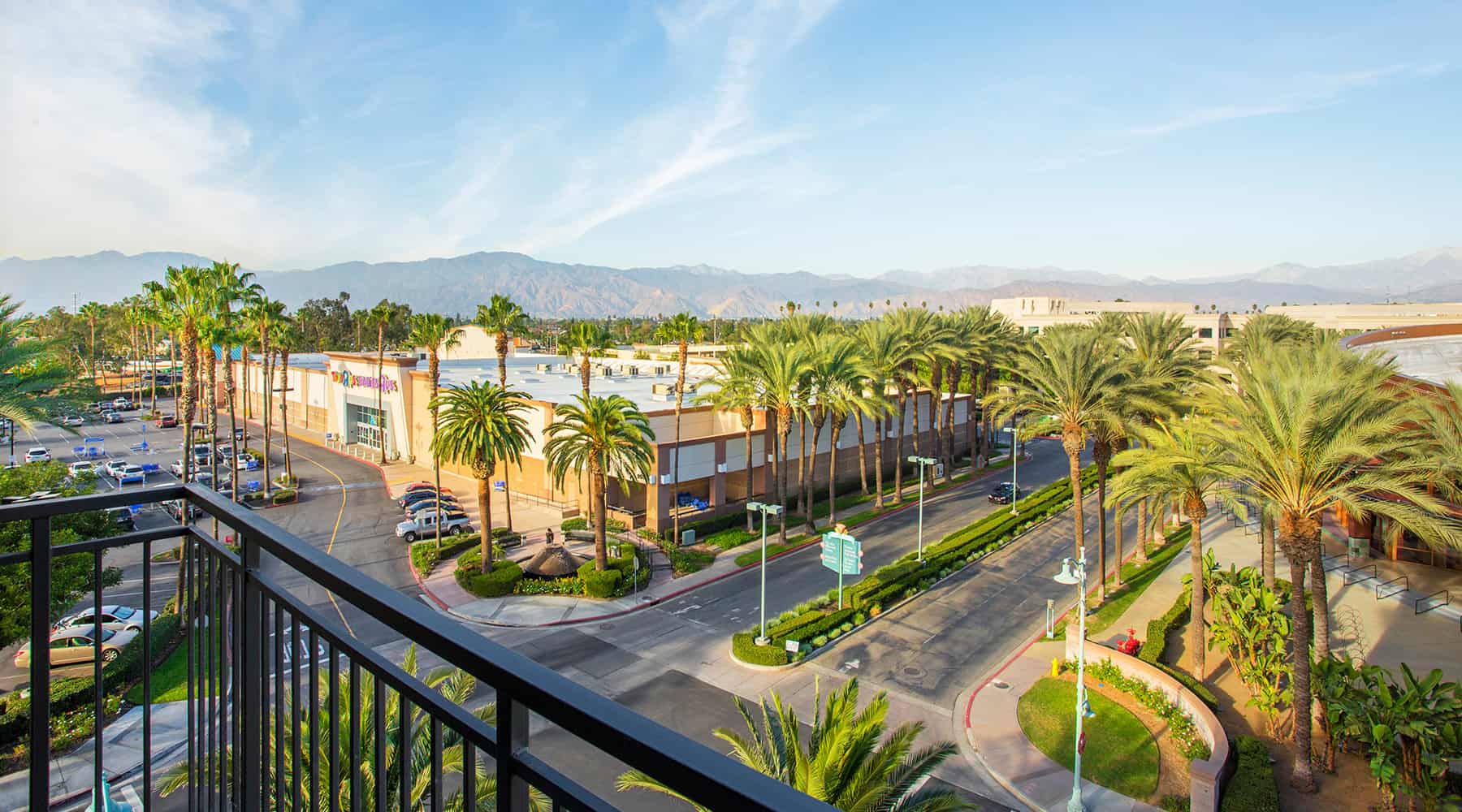 View from a balcony overlooking palm trees and a shopping center with mountains in the background.