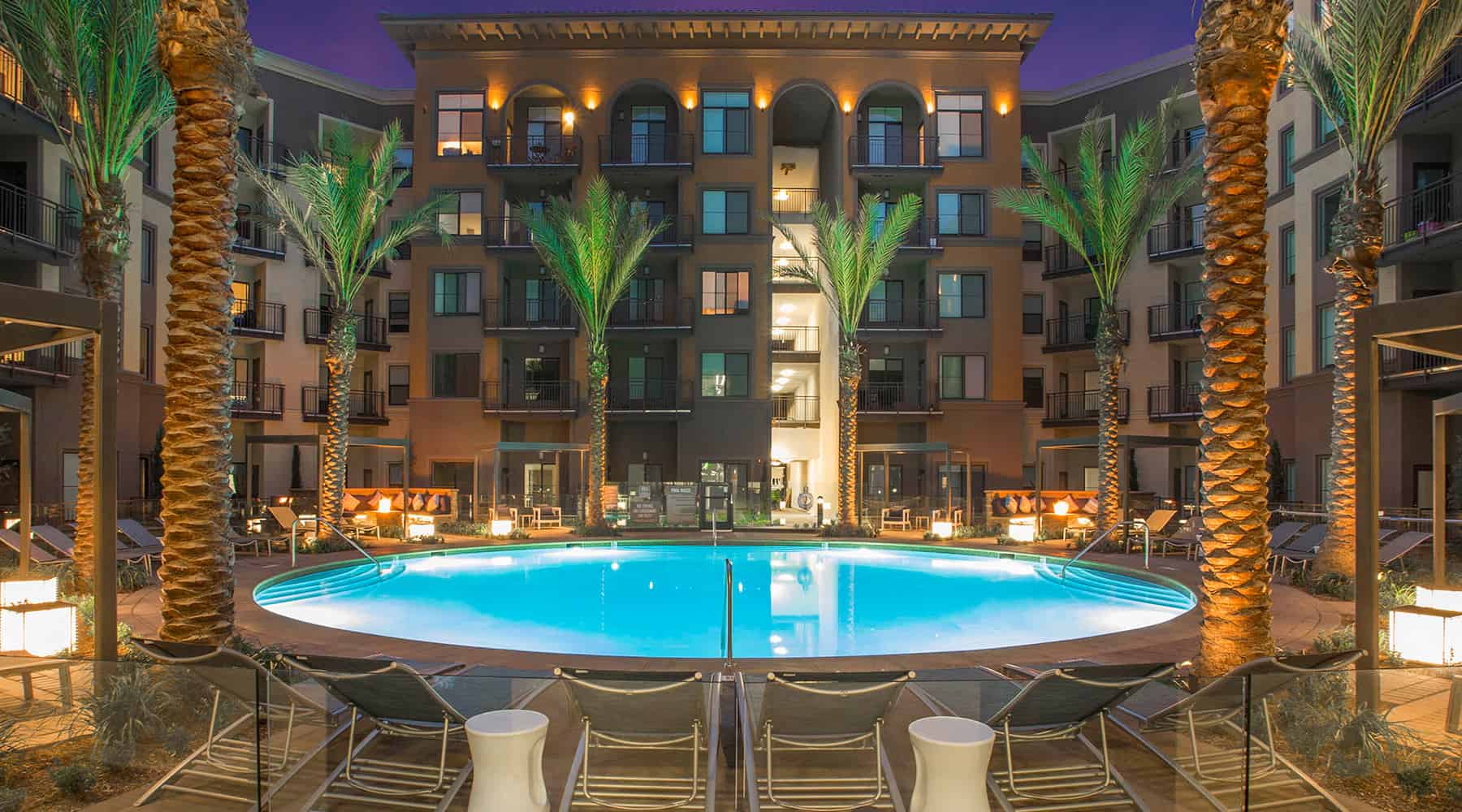 Evening view of a resort-style pool surrounded by palm trees and well-lit apartment buildings at Colony at the Lakes.
