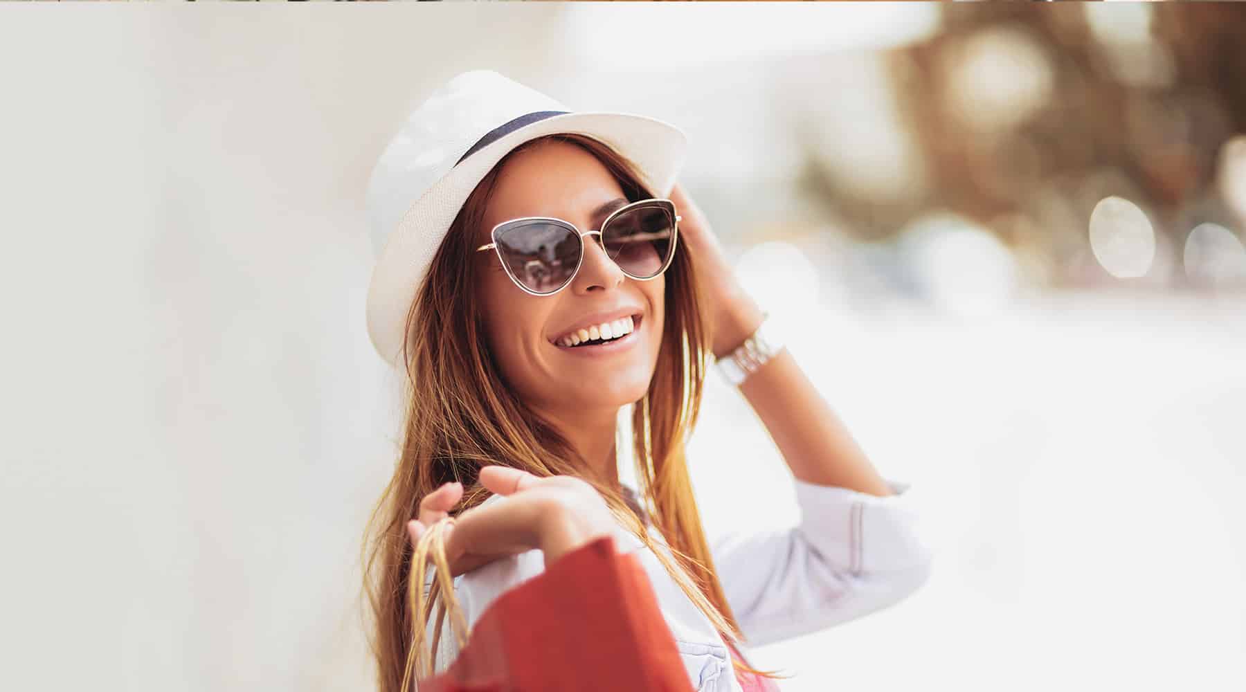 Woman smiling and posing with sunglasses and a white hat.