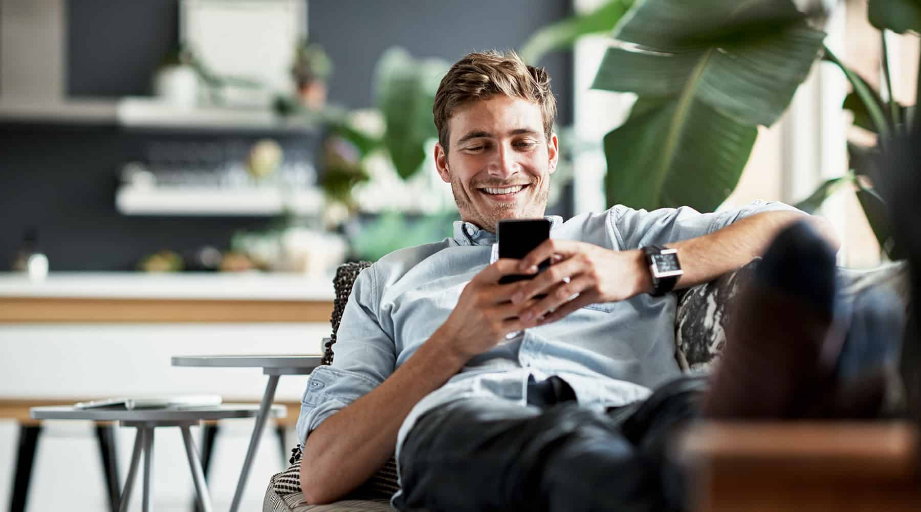 Man sitting on a couch, smiling while texting on his smartphone.