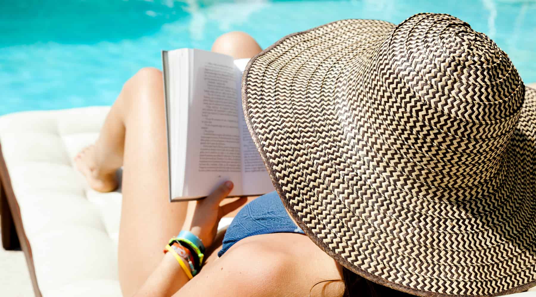 Person relaxing by the pool with a large sunhat and book in hand.